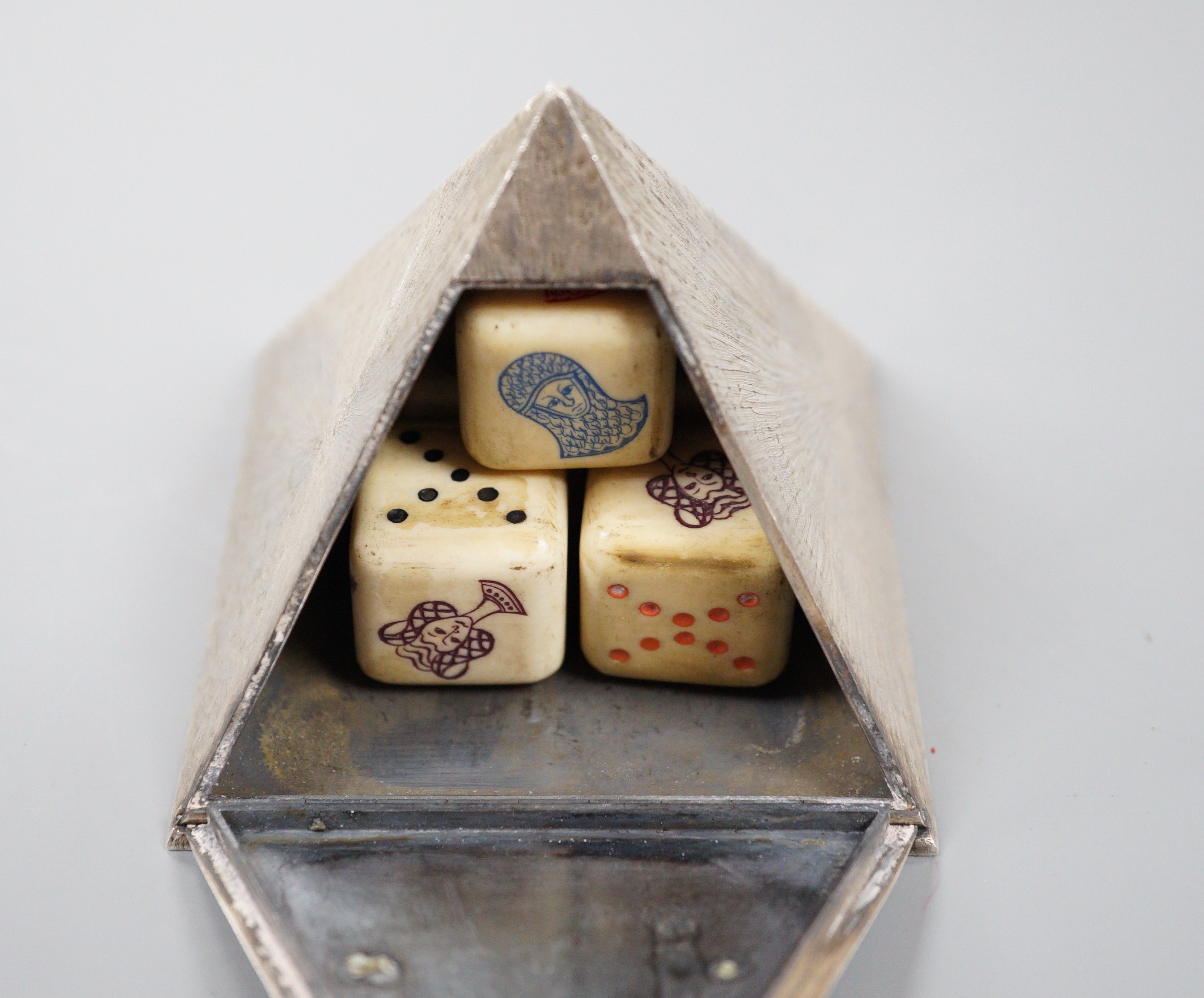 A textured white metal pyramid shaped box, the hinged panel decorated with Greek scene and containing five dice, height 62mm.